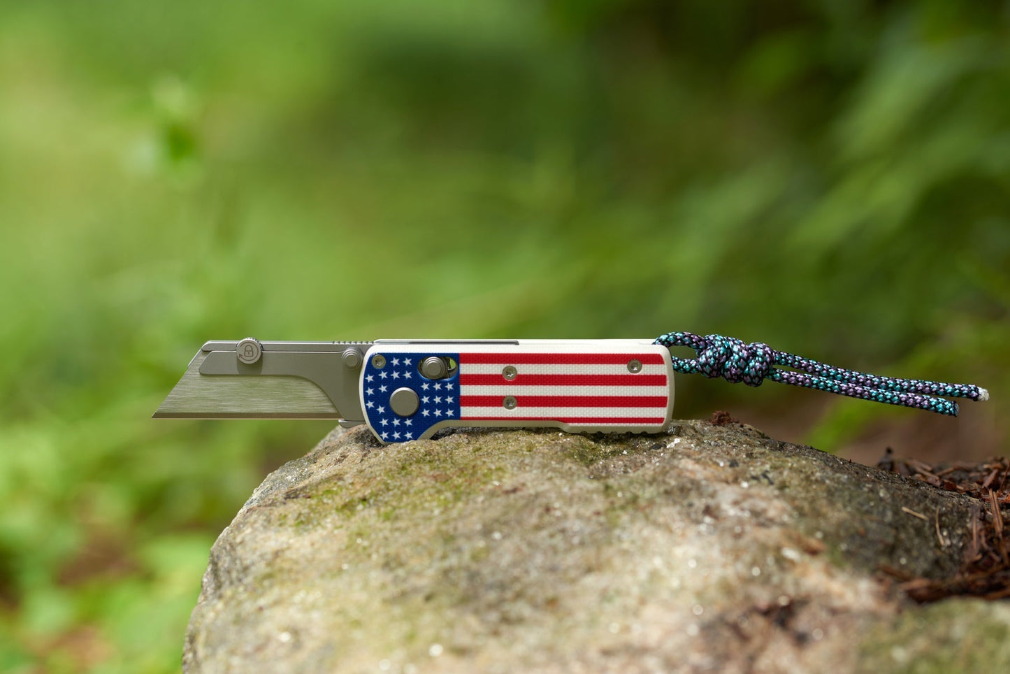 Oknife U1 Stars & Stripes open on a mossy rock outdoors showing the 2.23 inch replaceable SK5 utility blade and paracord lanyard