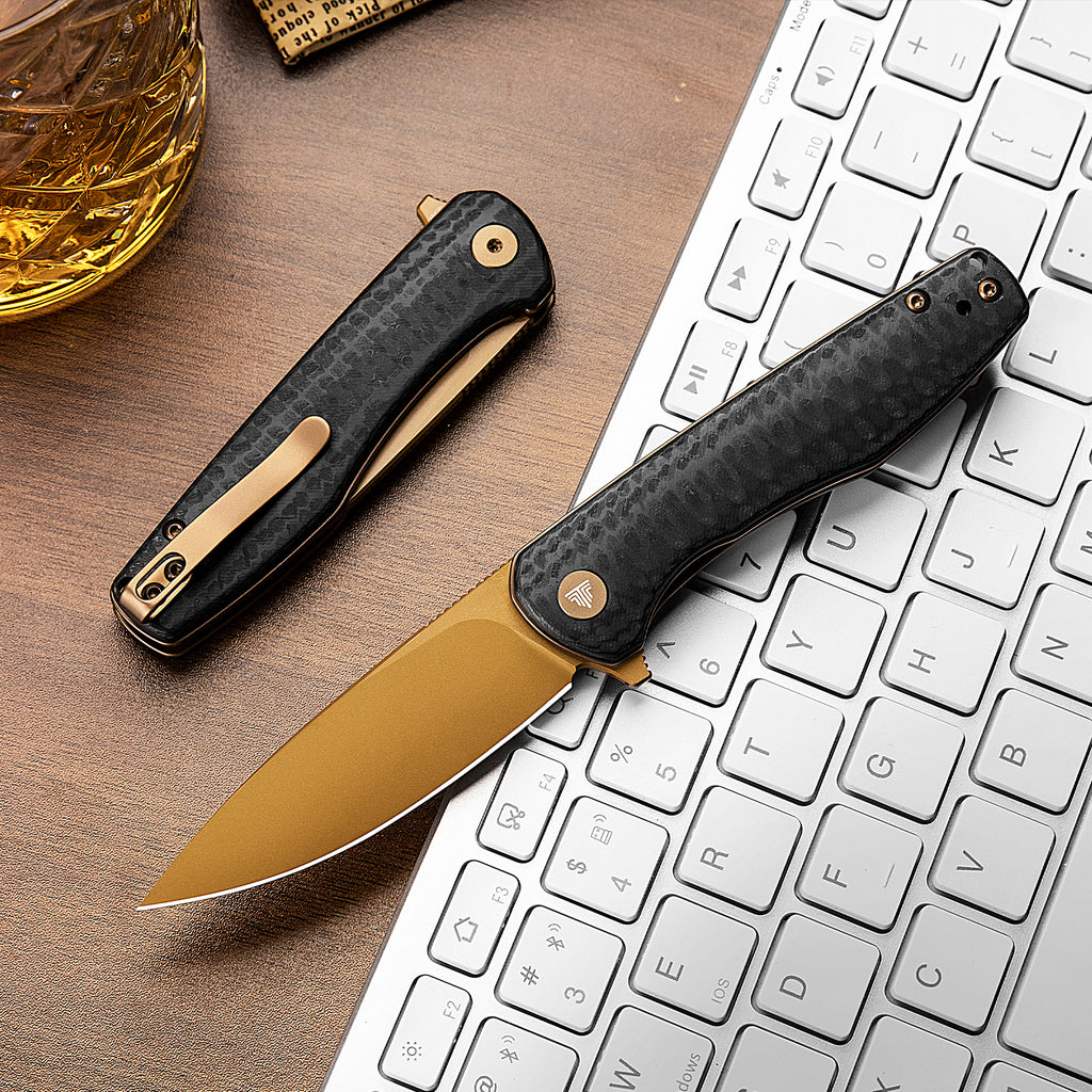 TRIVISA Hummingbird-CG folding knife with gold blade and carbon fiber handle on desk